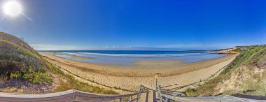 Geniş kumlu sahil ve berrak mavi gökyüzü olan sakin okyanus dalgaları, gündüz vakti Büyük Okyanus Yolu.