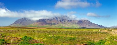 Geniş İzlanda manzarası. Kayalık dağlar kısmen bulutlarla ve yeşil alanlarla kaplı.