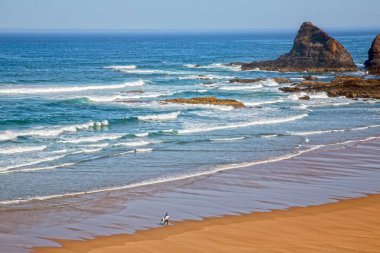 Dalgalar, Alentejo Portekiz kıyılarında yalnız bir sörfçü yürürken kumlu bir plaja yuvarlanıyor.