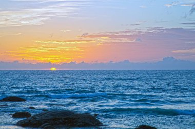 Phuket 'teki Kata Sahili üzerinde kayalık kıyı şeridi ve sakin okyanus dalgalarıyla altın gün batımı