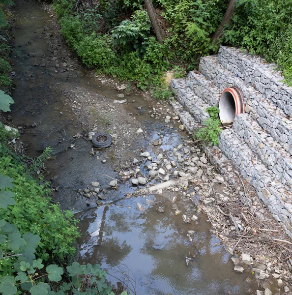 Dry riverbed. Lack of water in the tributary from the pipe protruding from the stone-armed and wired bank. Stones, an old tire, and other garbage are visible on the riverbed