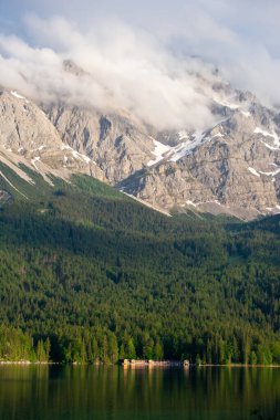 Almanya, Bavyera 'daki Eibsee Gölü' nde orman kıyısında küçük göl evleri ve kulübeleriyle, Zugspitze dağlarının altında kısmen bulutlarla kaplı. Manzaralı bir dağ manzarası.
