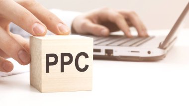 Arka planda ahşap küpler tutan bir adam PPC harfleri beyaz bir ofis masasında dizüstü bilgisayar.