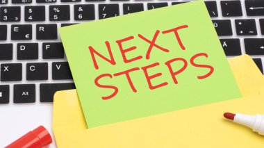 Yeşil ve sarı kartta kırmızı harflerle NEXT STEPS yaz. Çerçevede dizüstü bilgisayar klavyesi, kırmızı kalem, beyaz tahta masa var. iş ve finans kavramı.