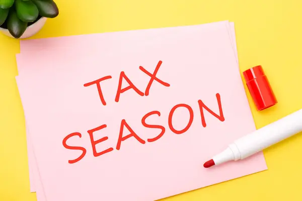 paper with TAX SEASON text stands on a yellow background with red marker, business concept