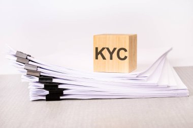 Bir yığın belgenin üzerinde KYC yazılı bir tahta blok. Gri masa, beyaz arkaplan