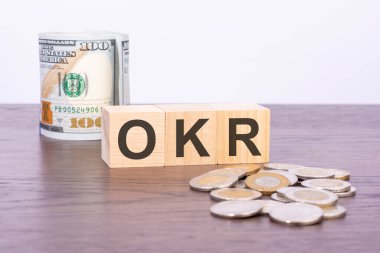 Kahverengi arka planda ABD doları banknotları üzerinde OKR (Hedefler ve Anahtar Sonuçlar) yazılı ahşap blokların üst görünümü. Vergi ve varil kavramı
