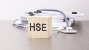 HSE - 'HSE - Sağlık ve Güvenlik Yöneticisi' gri bir arkaplanda steteskopun yanındaki tahta bloğa yazılmıştır