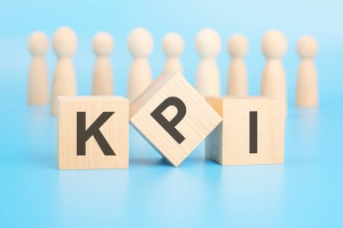 Önplanda bloklar olan kavramsal bir kelime oluşturmak - KPI (Anahtar Performans Göstergeleri). Bloklar, insanları sembolize eden ahşap figürlerin önünde mavi bir yüzey üzerinde yer alıyor..