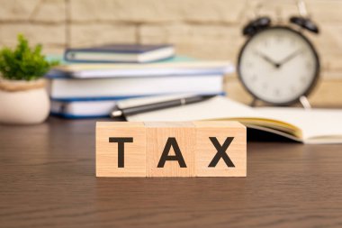 Ofis masasındaki ahşap küpün üzerine bir yığın ders kitabı, bir çalar saat ve arka planda saksı bitkisi yerleştirilmiş bir vergi mesajı..