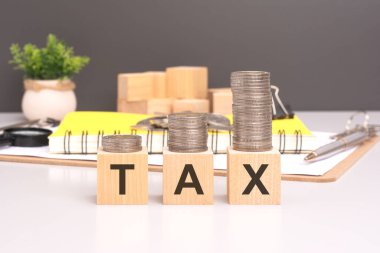 Tahta bloklara yığılmış madeni paralarla yazılmış vergi metinlerinin yakın çekimleri. Ofis masası arka planda. Ofis masasının arkasında sarı not defteri, kalem ve büyüteç var.