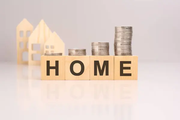 Close-up of coins on wooden blocks with the word home in focus. Wooden house models are blurred in the background. Business in the real estate sector.