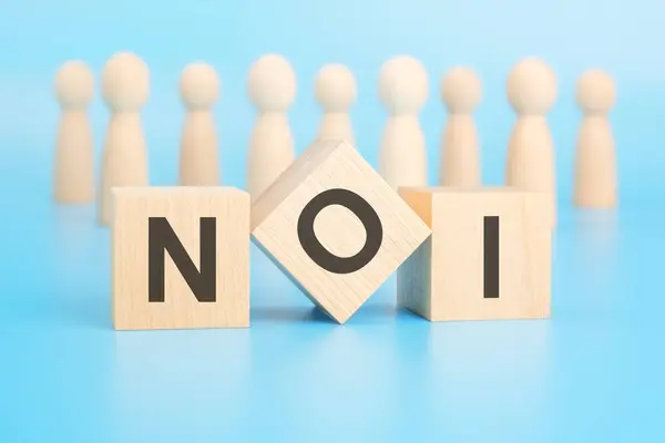 stock image forming a conceptual word with blocks in the foreground - NOI (Net Operating Income). the blocks are located on a blue surface in front of wooden figures symbolizing people.