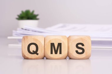 Ofis masasının üzerinde 'QMS' yazan tahta küpler, arka planda beyaz kağıt bir belge, QMS - Kalite Yönetim Sistemi 'nin kısaltması