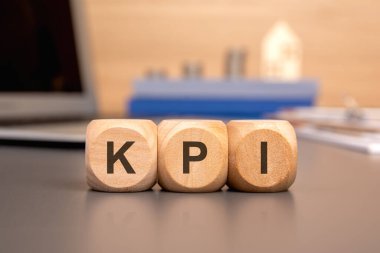 KPI 'yi tahta küplere, küpleri yansıtıcı bir yüzeye yaz. Arka planda iş bilgisayarı, madeni paralar ve model ev var.