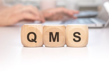 QMS kısaltmasını kodlamak için sıralanmış tahta küpler, arka plandaki dizüstü bilgisayarda bulanık ellerle yazıyorlar.