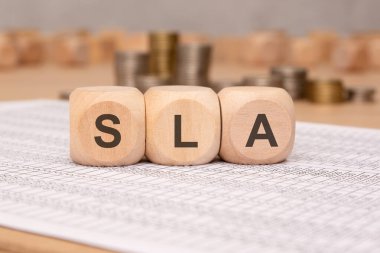 SLA kısaltması, finansal bir tabloya yerleştirilmiş ahşap bloklardan oluşur. Bulanık arkaplanda para yığınları hizmet düzeyi anlaşmalarını temsil eden profesyonel ve mali bir bağlam ekler