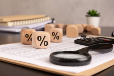 Bu resim, yüzde işareti ile işaretlenmiş tahta küplerle finansal analiz ve faiz oranı kavramlarını vurguluyor. Arka planda büyüteç, hesap makinesi ve ofis ögeleri hassasiyet ve veriye dayalı kararları vurgular.