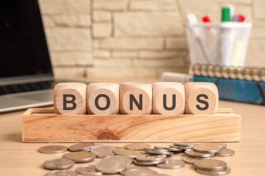 Görüntü finansal ödülleri ve teşvikleri gösteriyor. 'BONUS' kelimesini oluşturan tahta küpler dağınık madeni paralarla dolu bir masanın üzerine yerleştiriliyor, maaş primleri, çalışan teşvikleri ve iş gelirlerini simgeliyor..