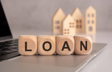 İmaj ev kredileri, ipotekler ve emlak finansmanını temsil ediyor. Kredi kelimesini oluşturan tahta küpler dizüstü bilgisayara yerleştiriliyor ve arka planda minyatür ahşap evler konut finansmanını simgeliyor..