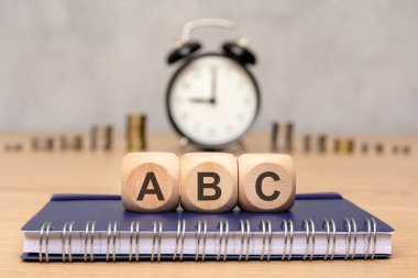 'ABC' li ahşap küpler spiral bir deftere yerleştirilmiş, alarm saati ve arka planda birikmiş paralar, öğrenme, işletme, zaman yönetimi, finansal okur yazarlık ve eğitim kavramlarını temsil ediyor.