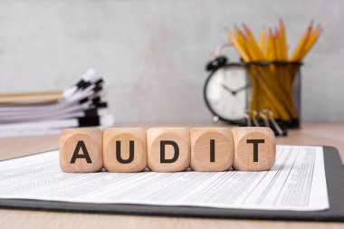 Arkaplanda ofis malzemelerinin bulunduğu mali raporlara 'AUDIT' sözcüğü konan tahta bloklar. Mali denetimi, muhasebeyi, uyumu, vergi incelemesini ve kurumsal değerlendirmeyi simgeliyor