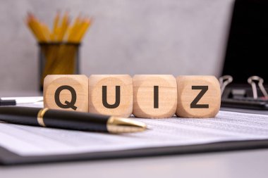 Bilgi, test, değerlendirme ve öğrenmeyi temsil eden finansal belgelere 'QUIZ' sözcüğünü yerleştiren tahta küpleri. Arkaplanda ofis malzemeleri akademik veya mesleki bir ortamı vurgular