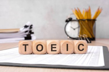 wooden blocks forming the word 'TOEIC' placed on reports with office supplies in the background. Symbolizing Test Of English For International Communication clipart