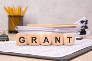 Fon, burs ve finansal desteği sembolize eden, arka planda ofis kırtasiyesiyle birlikte GRANT sözcüğünün sergilendiği tahta küpler