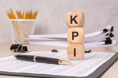 Çalışma raporları, kalem ve ofis malzemelerinin olduğu bir masada KPI sergilenen tahta küpleri. Organizasyonlarda temel performans göstergeleri, iş ölçümleri ve performans ölçümlerini temsil eder