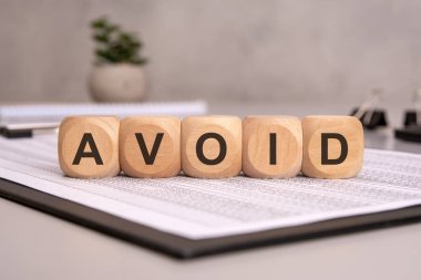 AVOID kelimesinin hecelendiği küpler finansal belgelere yerleştiriliyor. Kalemler, kağıtlar ve arka planda küçük yeşil bir bitki bulunan bulanık bir ofis masası profesyonel ve karar verici bir atmosfer oluşturur..
