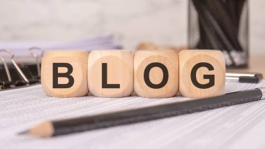 Ofis malzemeleriyle dolu bir masada BLOG kelimesini oluşturan dört tahta küp, profesyonel bir çalışma alanında içerik oluşturma, yazma ve dijital iletişimi vurguluyor