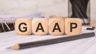 Genel Kabullenen Muhasebe İlkelerini sembolize eden, GAAP ile finansal bir hesap tablosu üzerinde ahşap küplerin yakınlaştırılması. Ofis malzemeleri kalem ve klips gibi arka planda muhasebe ve finans temasını vurguluyor.