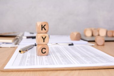 KYC 'nin dikey olarak dizilmiş tahta küpleri, arka planda kalemler ve ofis malzemeleriyle finansal belgelerinin üzerine yerleştirilmiş, Müşterinin Maliye ve Uyum sürecini temsil eden,