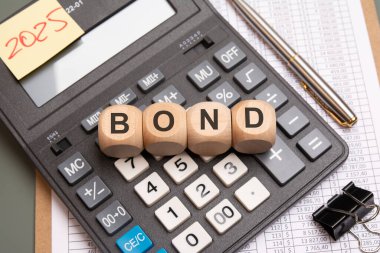 Hesap makinesinde BOND sözcüğünü oluşturan ahşap küplerin yakın plan görüntüleri. Finansal hesap tabloları, bir kalem, bir cilt klipsi ve 2025 mali temalı bir kompozisyon olan yapışkan bir not.