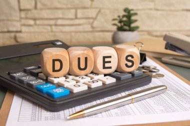DUES kelimesi, mali belgeler, madeni paralar, bir kalem ve ofis eşyalarıyla çevrili bir hesap makinesine yerleştirilmiş ahşap küplerle yazılır. Ödemeler, borçlar ve mali yükümlülükler için kavramsal imaj.