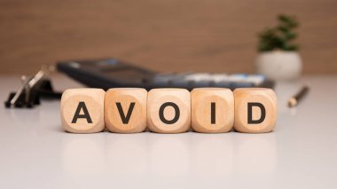 Modern ofis masasının üzerinde AVOID sözcüğünün yazılı olduğu tahta zar. Hesap makinesi ve klasör klipleri gibi iş aletleri içeren bulanık bir arkaplan. Sunumlar, bloglar ve eğitim içeriği için mükemmel.