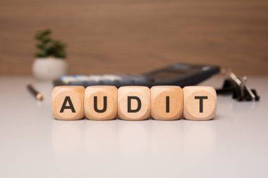 Arkaplanda bir kalem, belge ve hesap makinesi ile beyaz bir masa üzerinde AUDIT sözcüğünü oluşturmak için ahşap küpleri hizalanmış.