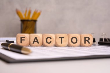 Ofis malzemeleri, kalemler ve belgelerle dolu bir masanın üzerindeki tahta bloklarla yazılan kelime FACTOR..