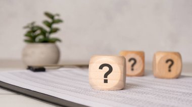 Tahtaya basılı hesap tablosu ile birlikte soru işaretleri konulmuş üç tahta küp. Arka planda hafif ışık ve bulanık bitkiyle en az ofis sahnesi. Şüphe ya da soruşturma kavramı.
