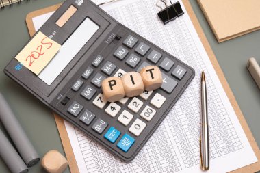 Finansal hesap tablosu, kalem, cilt klipsi ve 2025 notuyla çevrili bir hesap makinesinde PIT 'yi simgeleyen tahta küpler, vergi, finans ve kişisel gelir kavramını simgeliyor.