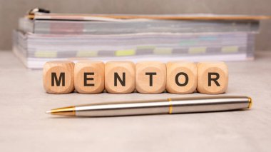 Ahşap blokların önü beton bir masanın üzerine yerleştirilmiş akıl hocasını oluşturuyor. Gösterişli metalik bir kalem ön planda yatarken, renkli renkli raporlar arkada bulanık duruyor, koçluğu, liderliği ve bilgiyi paylaşmayı sembolize ediyor.