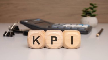Üç tahta blok beyaz ofis masasında KPI yazıyor. arka planda performans ve iş ölçümlerini sembolize eden bir hesap makinesi, cilt klipleri, kalem ve küçük bir bitki görünür.
