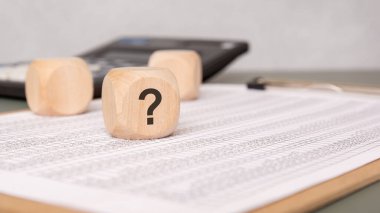 Finansal hesap tablosunda soru işareti olan tahta küp, arka planda bulanık hesap makinesi ve ofis malzemeleri. Şüphe, analiz ve mali kararlar kavramı