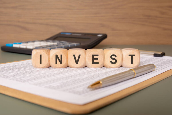 Концептуальная фотография инвестиций и финансов: деревянные кубики со словом INVEST, размещенные на финансовых отчетах, с ручкой и калькулятором на заднем плане.
