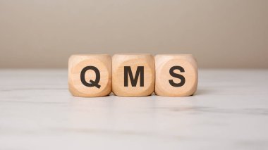 Hafif arkaplandaki tahta bloklara QMS harfleri, iş dünyasındaki kalite yönetim sistemini sembolize ediyor