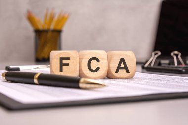 FCA kısaltması, iş masasında bulunan tahta küplerle, kağıt, kalem ve kalemlerle yazılıyor.
