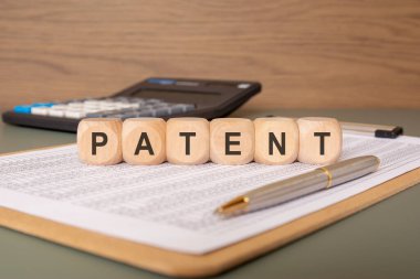 Ahşap blokların yakınlaştırılması PATENT sözcüğünü finansal bir tabloya yerleştiriyor. Arka planda, fikri mülkiyet ve yenilik finansmanını temsil eden bir kalem ve bir hesap makinesi bulunmaktadır..