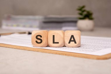 Üzerinde SLA harfleri olan üç tahta blok basılı hesap tabloları olan bir panoda duruyor. Arka planda, bulanık belgeler ve saksı bitkisi bir iş ortamı oluşturuyor. SLA, Hizmet Seviyesi Anlaşması anlamına geliyor.