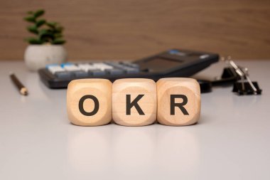 OKR (Hedefler ve Anahtar Sonuçları) kısaltmalı üç tahta küp bir ofis ortamında bir masanın üzerine yerleştirilmiş.